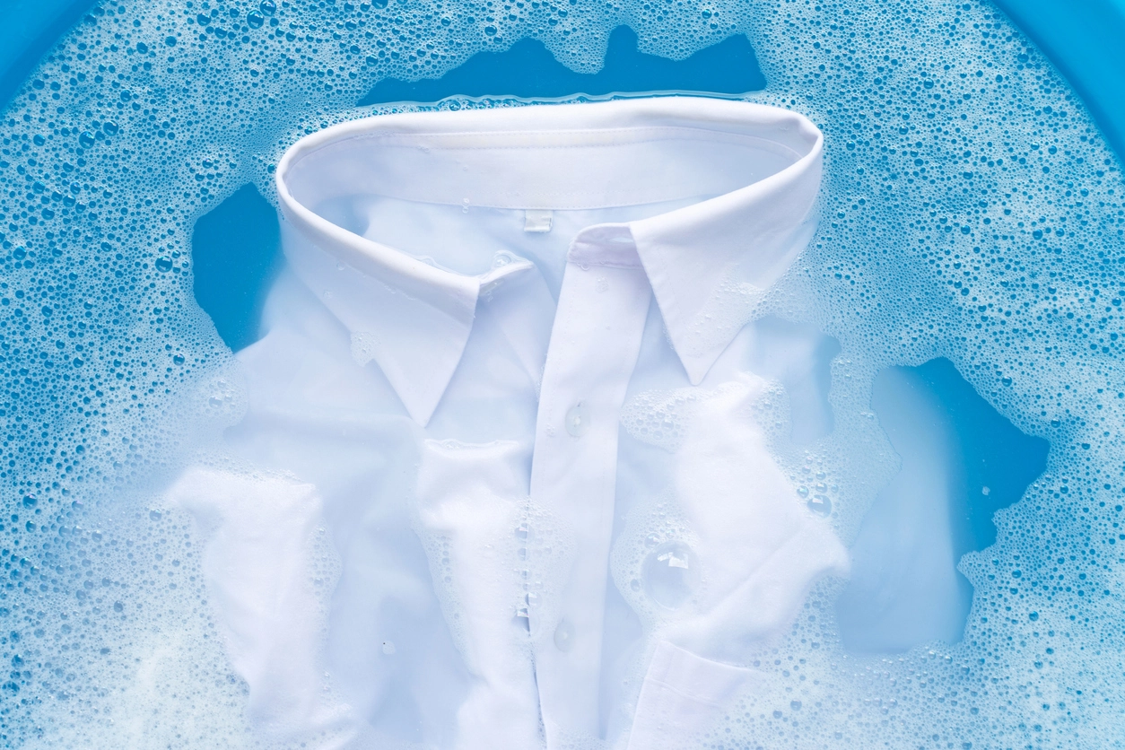 A white dress shirt is soaking in soapy water, surrounded by bubbles in a blue basin.